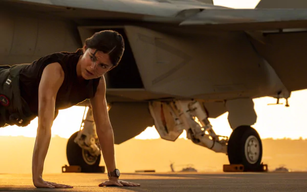 Monica Barbaro from Top Gun: Maverick performing push-ups on the tarmac near a jet, captured in an HD movie wallpaper background.