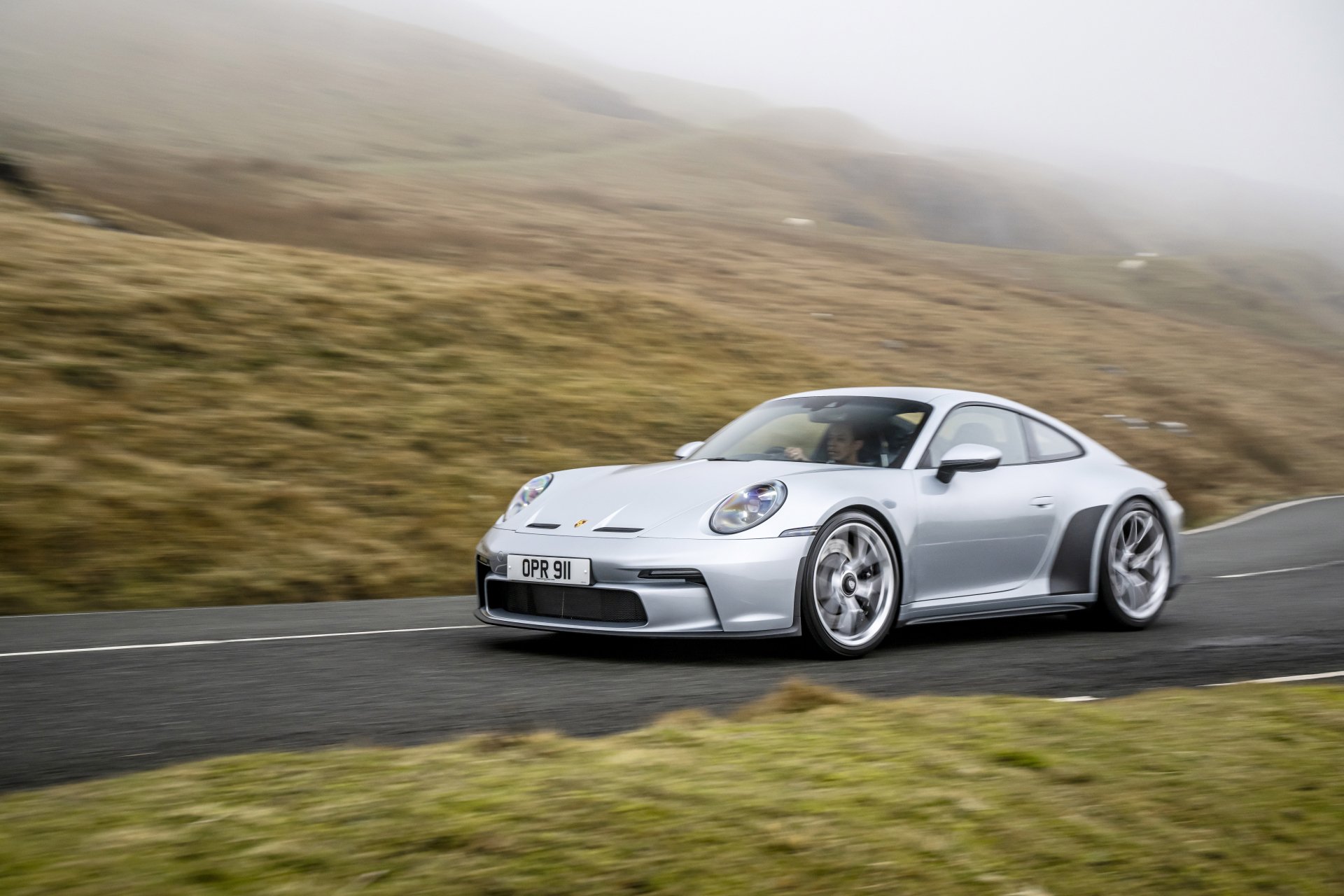 Silver Porsche 911 GT3 driving on a winding country road through misty hills, 8K Ultra HD PC desktop wallpaper/background.