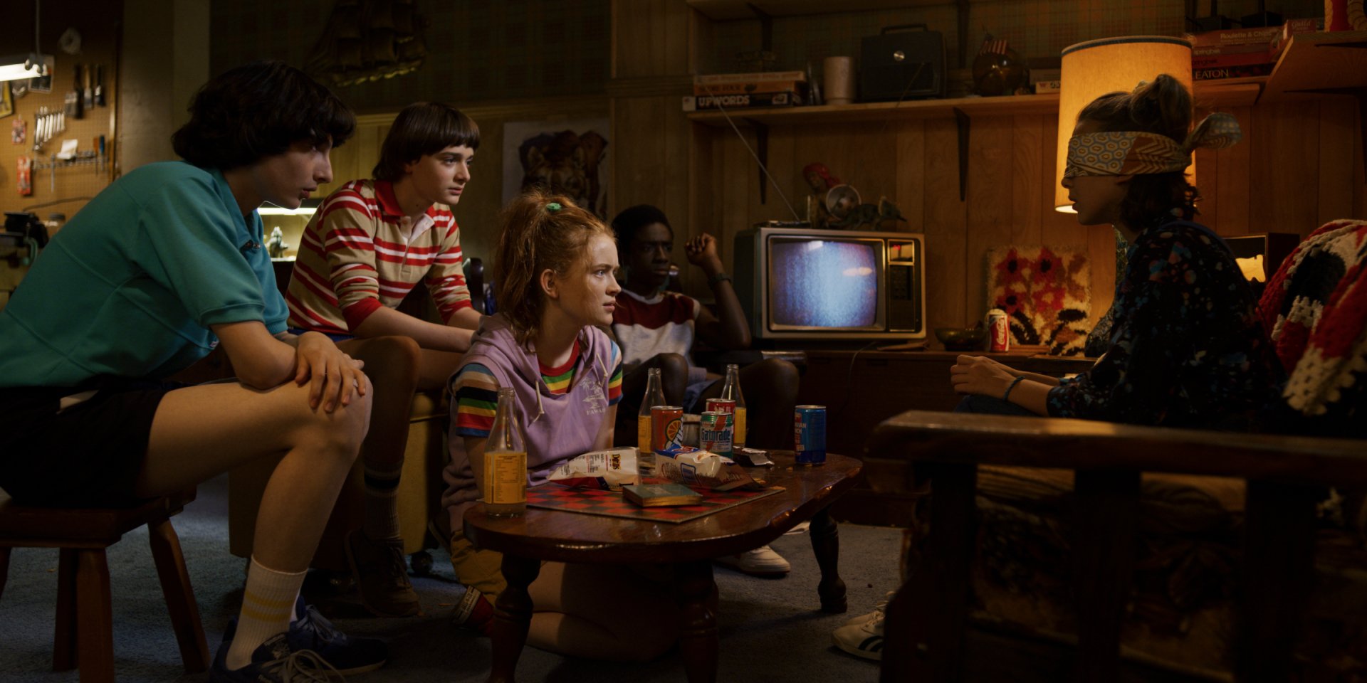 2K Quad HD PC desktop wallpaper: TV show Stranger Things scene — teens in a dim basement around a small TV, snacks on the coffee table, one girl blindfolded.