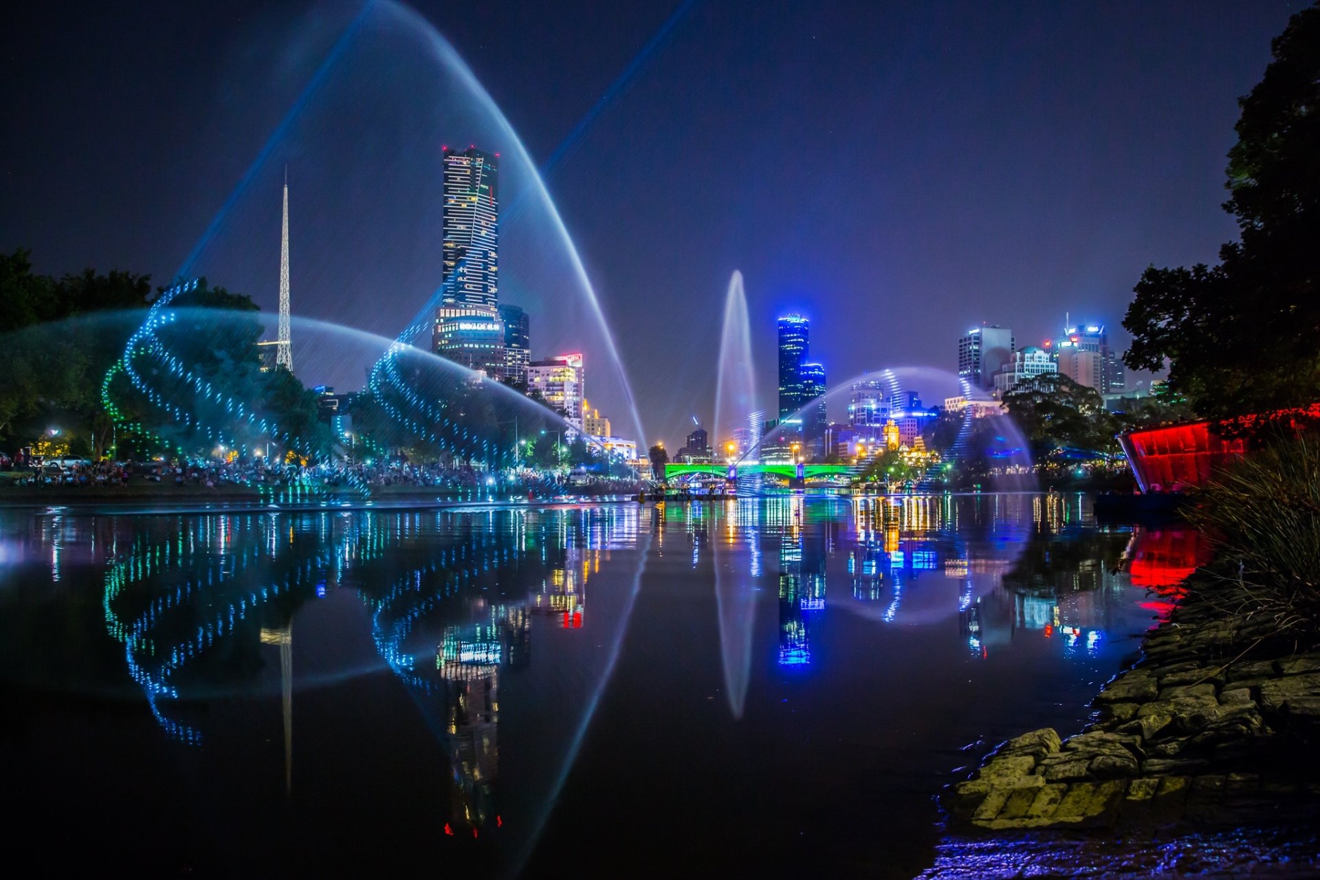 Nighttime view of Melbourne's city skyline and light reflections over calm water, showcasing vibrant man-made illumination and urban architecture.