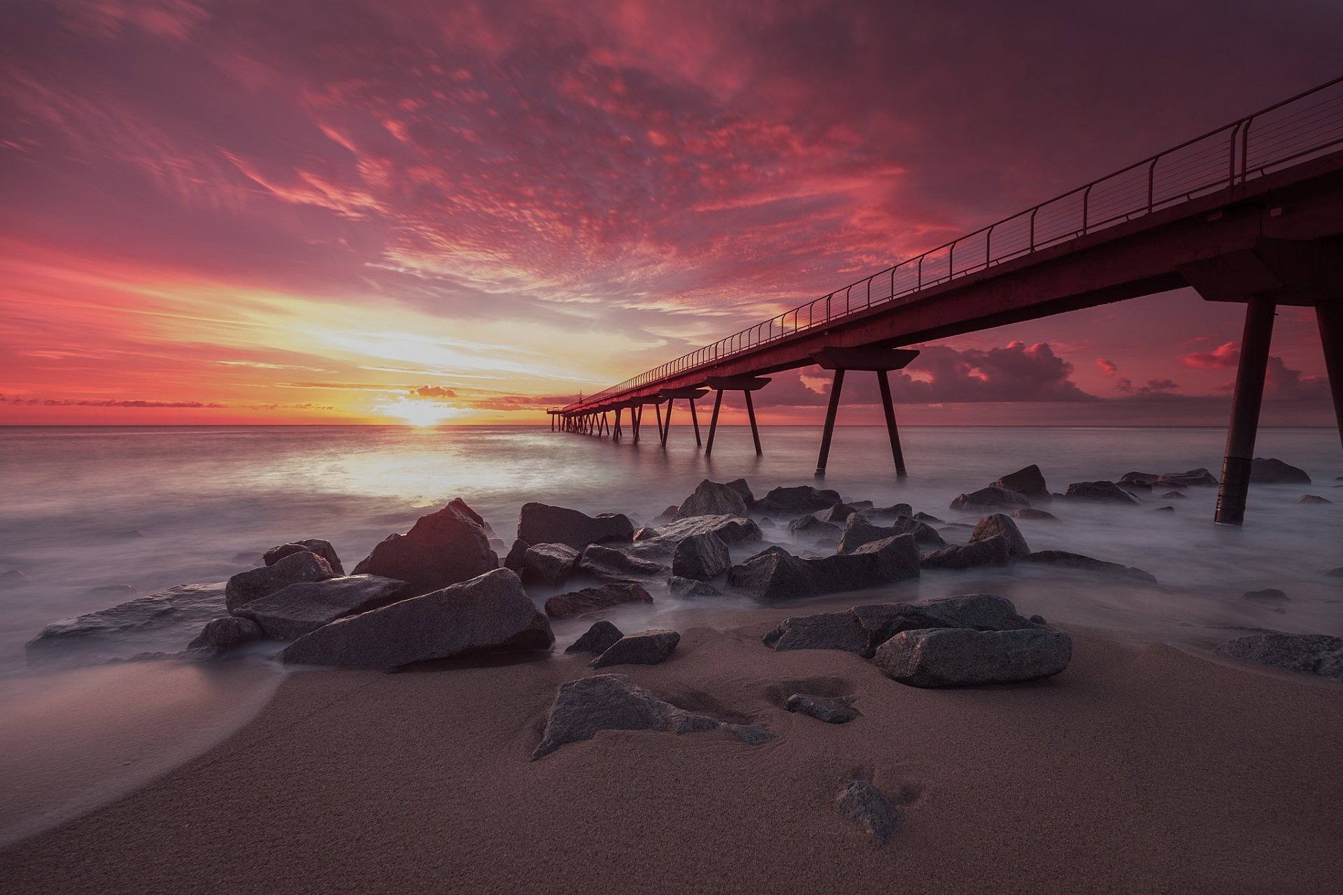HD PC desktop wallpaper: sunset horizon with a long man-made pier stretching over a rocky shore into a calm, colorful sea.