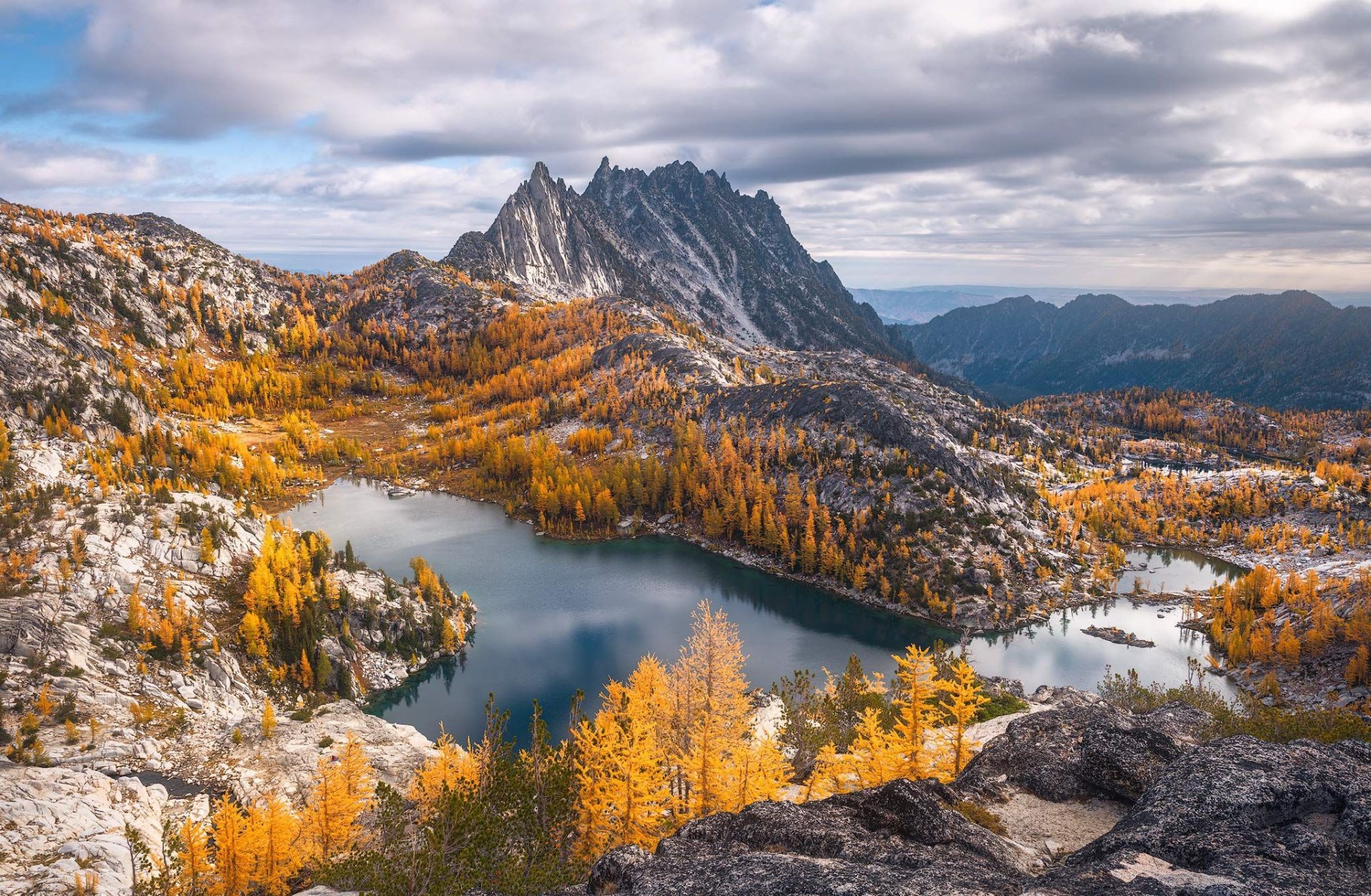 HD PC desktop wallpaper/background — alpine nature scene with jagged peaks and golden larches encircling a clear mountain lake beneath a dramatic cloudy sky.