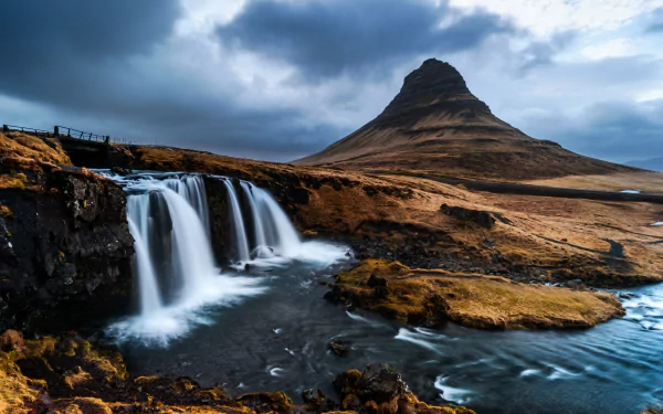A 4K Ultra HD image of Kirkjufoss waterfall flowing beside Kirkjufell mountain, showcasing stunning natural Icelandic landscape under a moody sky.
