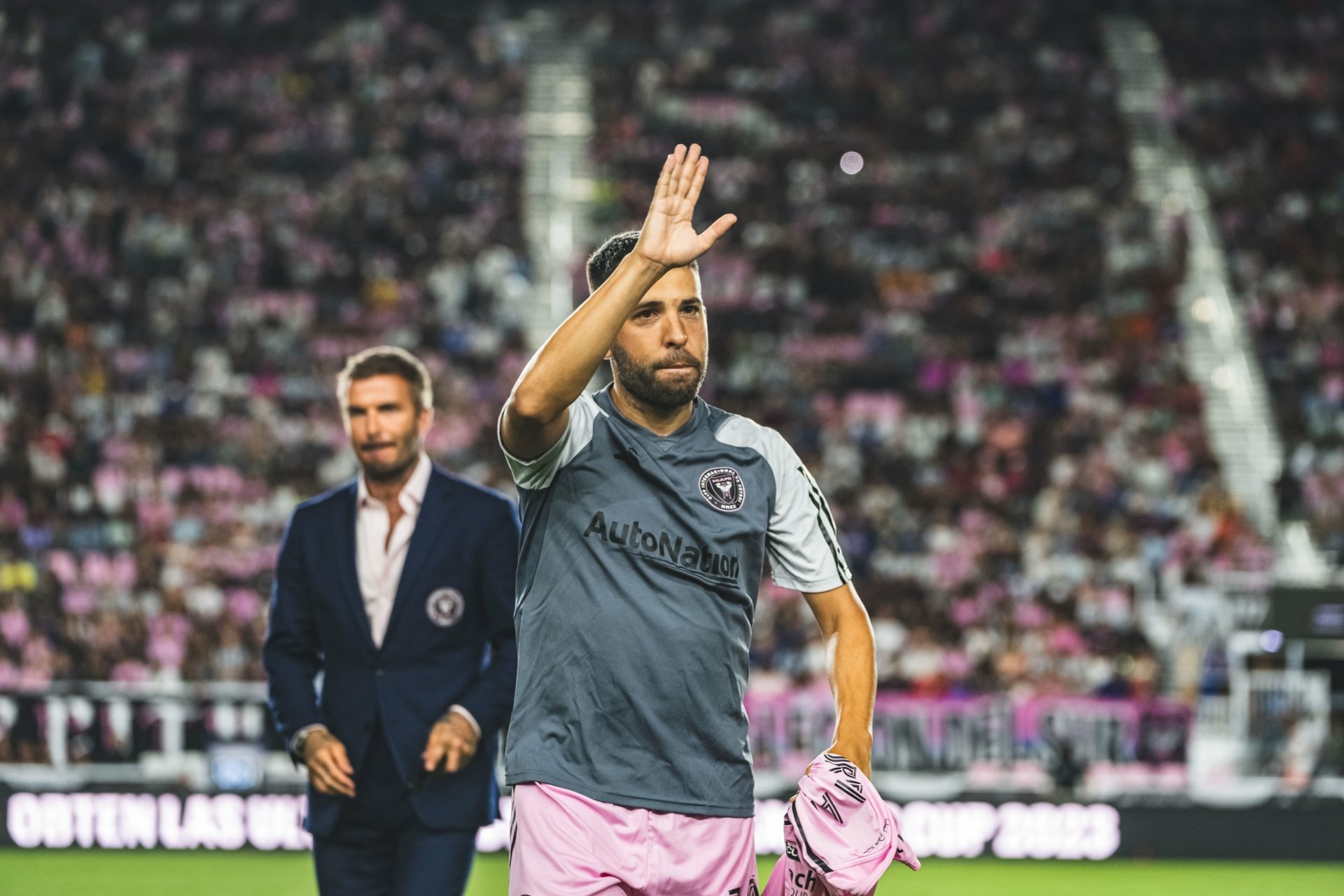 HD desktop wallpaper featuring Jordi Alba of Inter Miami CF waving on the field in a gray and pink uniform, with a blurred coach in the background.