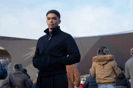 A character from the TV show Secret Invasion stands confidently in front of a modern structure, surrounded by a crowd, set against a cloudy sky. HD desktop wallpaper.