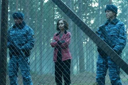 A tense scene from the TV show Secret Invasion, featuring a character in a pink jacket standing between two armed guards at a fence in a misty forest backdrop. HD wallpaper.