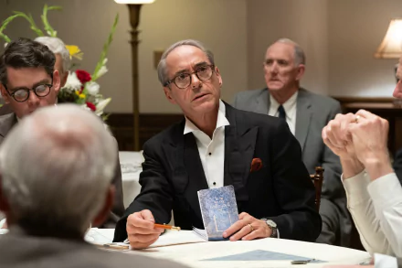 HD wallpaper featuring a pensive man in a suit sitting at a conference table, evocative of themes associated with Oppenheimer, portrayed by actor resembling Robert Downey Jr.