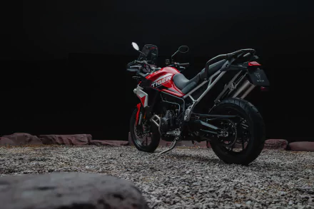 A vibrant Triumph Tiger 900 GT Aragón sits on gravel, showcasing its sleek design and bold red accents against a dark backdrop. This HD desktop wallpaper captures the essence of adventure.