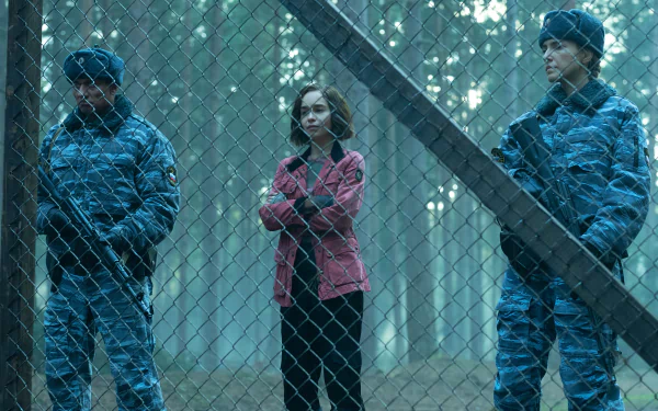 A tense scene from the TV show Secret Invasion, featuring a character in a pink jacket standing between two armed guards at a fence in a misty forest backdrop. HD wallpaper.