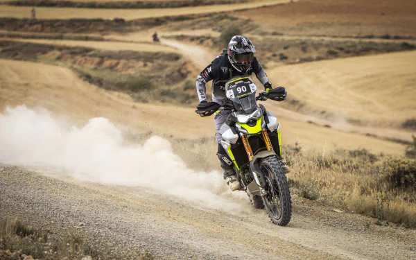 A rider skillfully maneuvers a Triumph Tiger 900 Rally Pro on a dirt path, kicking up dust against a breathtaking landscape, captured as an HD desktop wallpaper.