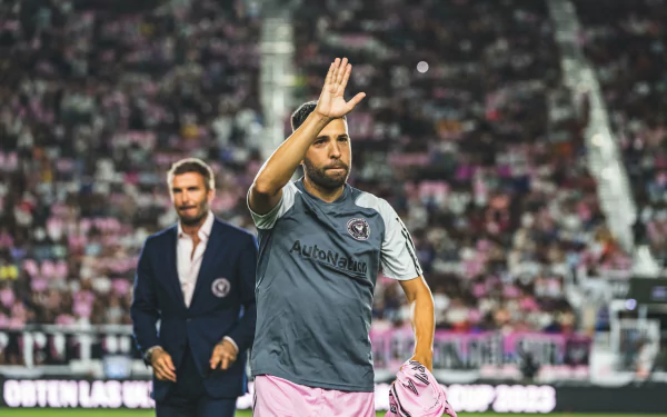 HD desktop wallpaper featuring Jordi Alba of Inter Miami CF waving on the field in a gray and pink uniform, with a blurred coach in the background.