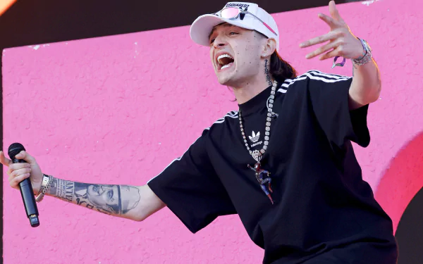 Performer gesturing enthusiastically on stage with a microphone, set against a vivid pink background, tagged with Peso Pluma - HD desktop wallpaper.