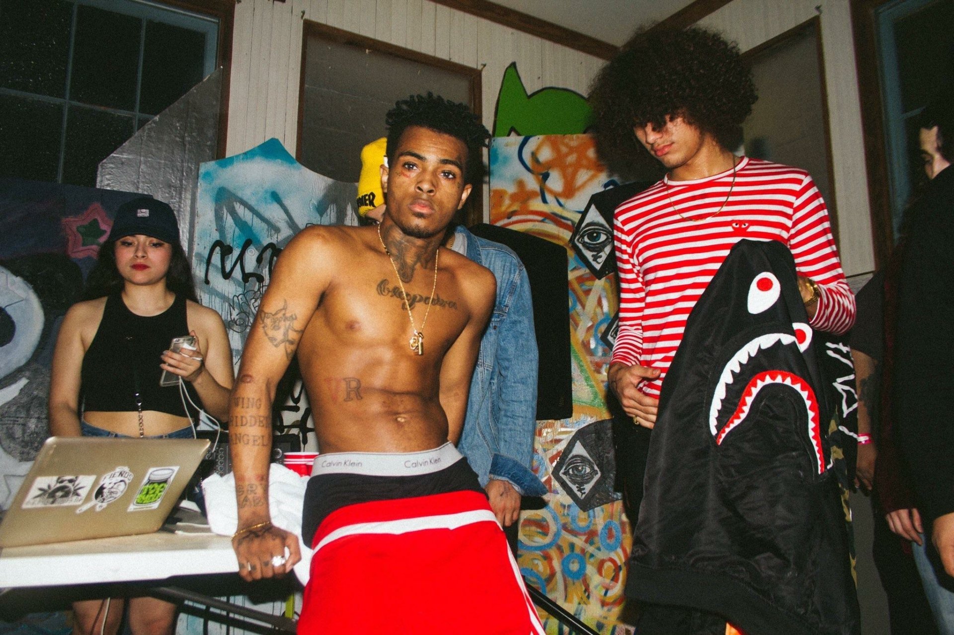 HD PC desktop wallpaper featuring XXXTentacion and friends in a casual indoor setting with graffiti-covered walls, capturing a raw music vibe.