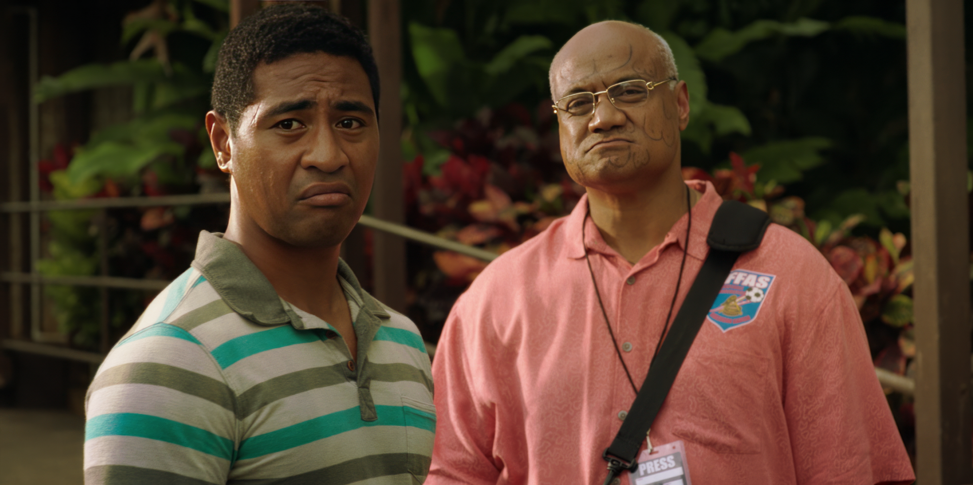 HD wallpaper featuring two men from the film 'Next Goal Wins (2023),' one with a quizzical expression and the other looking stern, set against a backdrop with lush greenery.