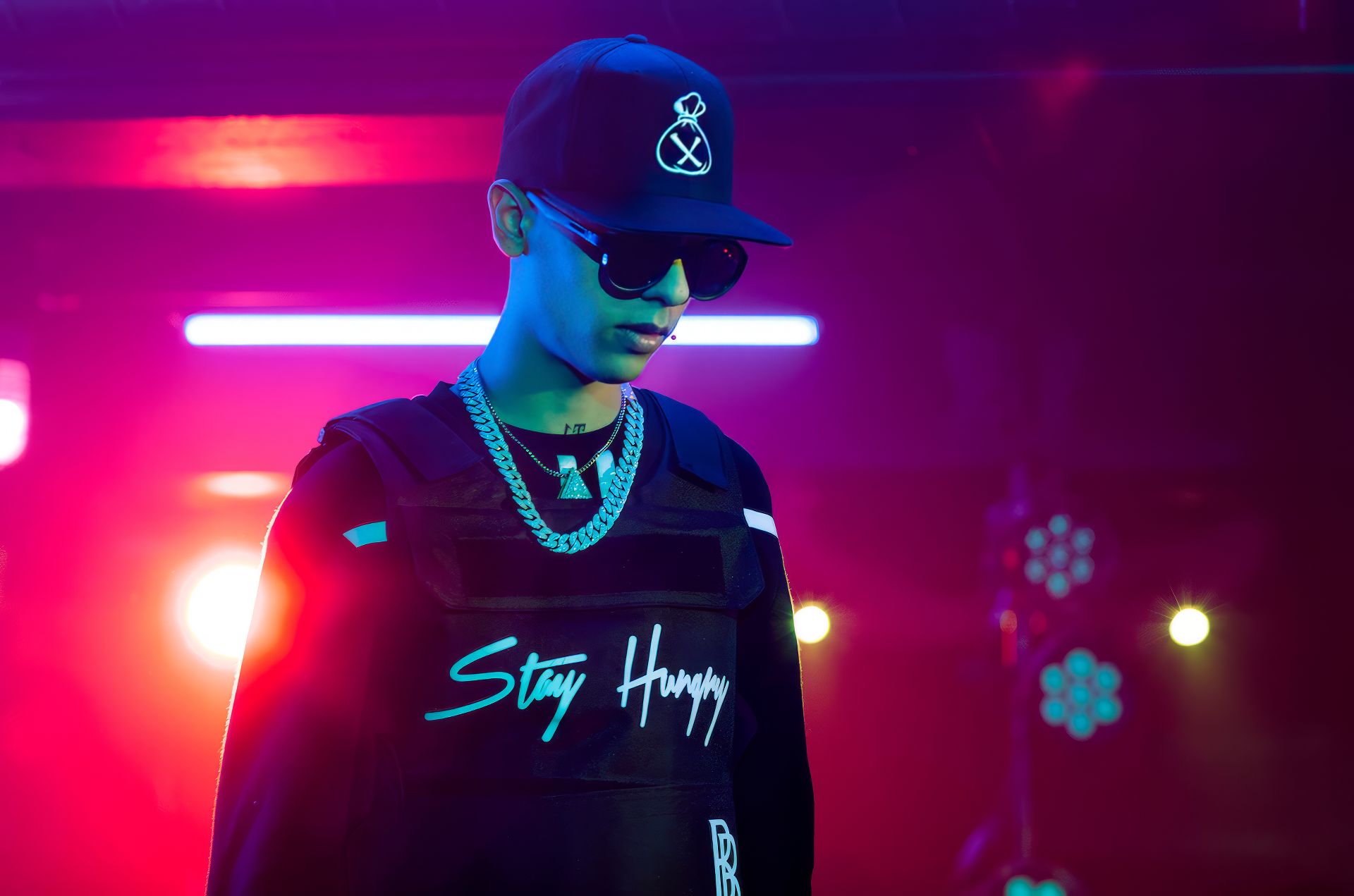 HD desktop wallpaper featuring a stylized image of a person with sunglasses and a baseball cap standing against a vibrant blue and purple neon light background, emblazoned with the slogan 'Stay Humble.'