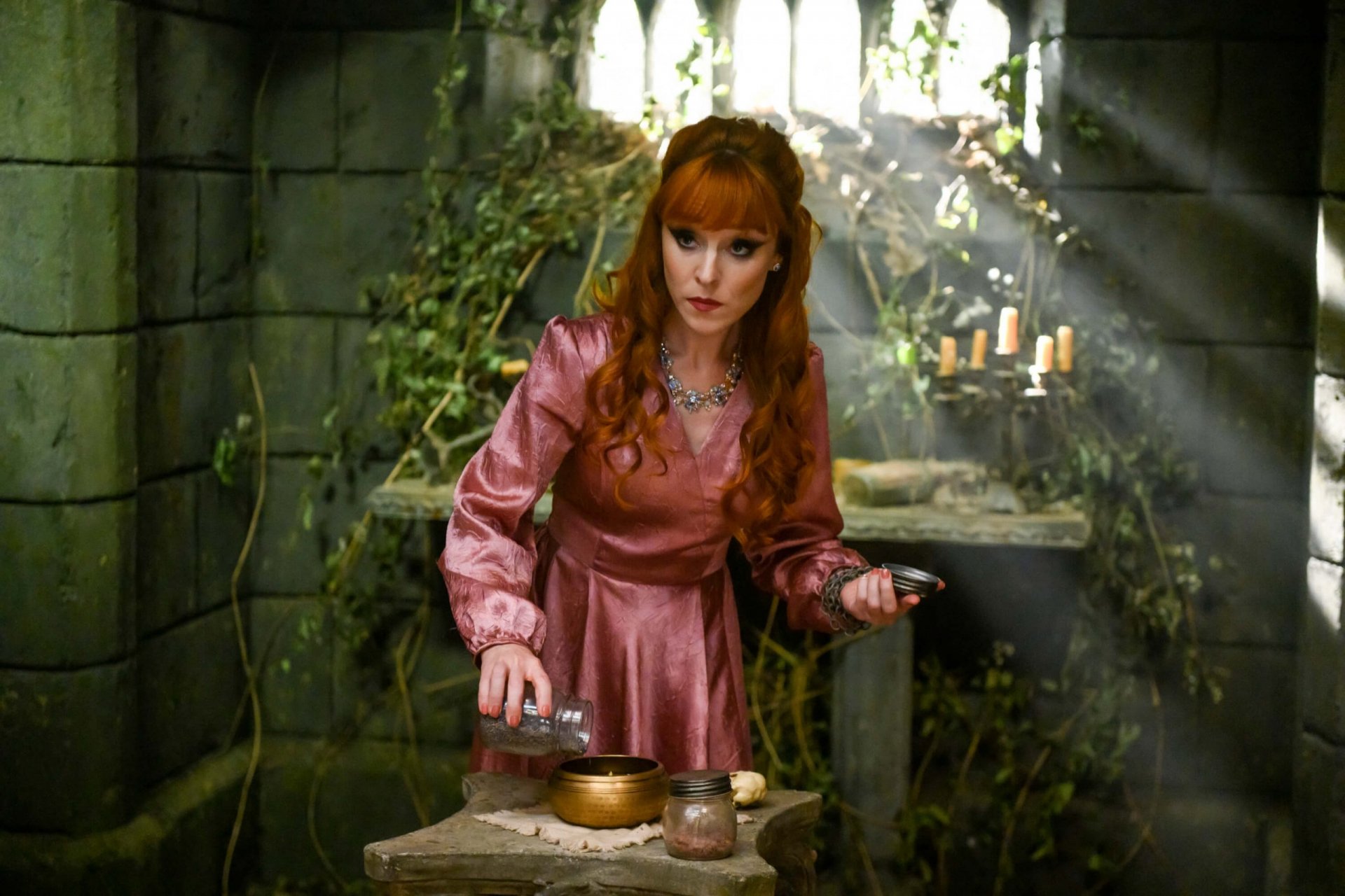 HD PC desktop wallpaper featuring a red-haired woman in a pink dress from the TV show Supernatural, standing in a dimly lit, mystical stone room with candles and greenery.