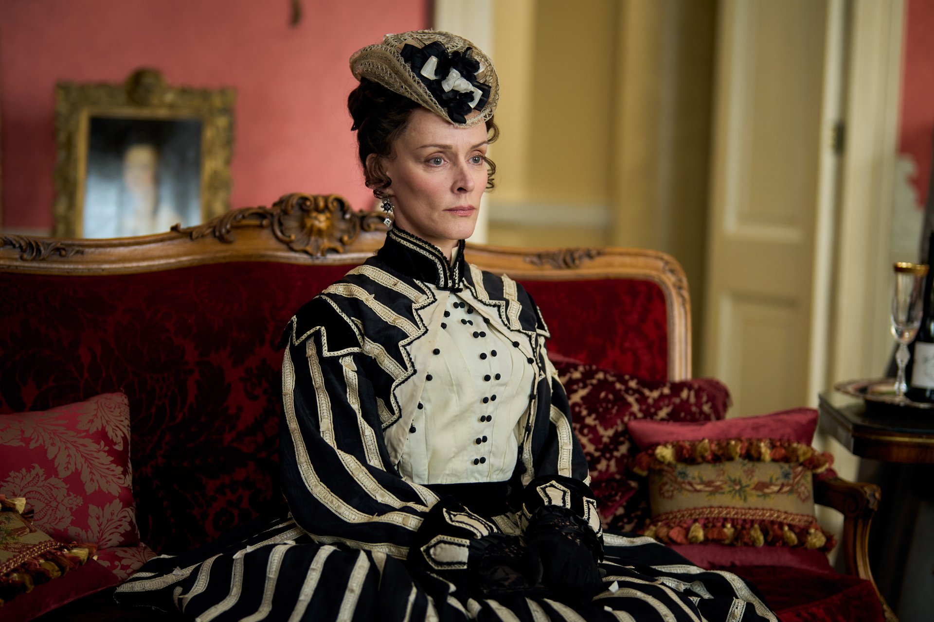 Elegant character from Belgravia: The Next Chapter dressed in Victorian attire sitting on an antique sofa with luxurious red background – HD desktop wallpaper.