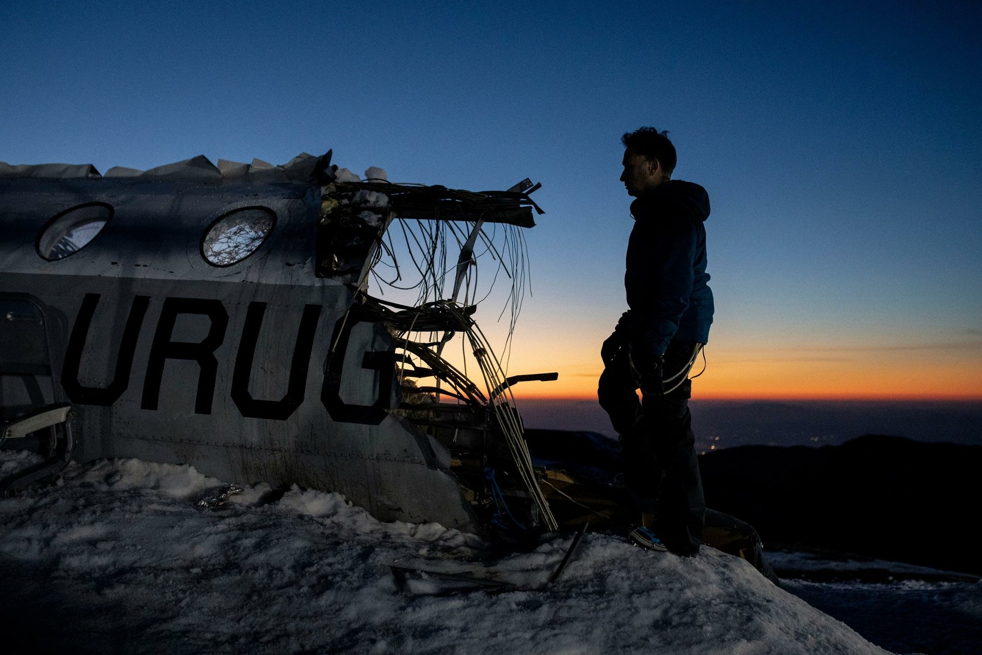 A person standing beside a wrecked airplane with the word 'URUG' at dusk - HD wallpaper inspired by the movie Society of the Snow.
