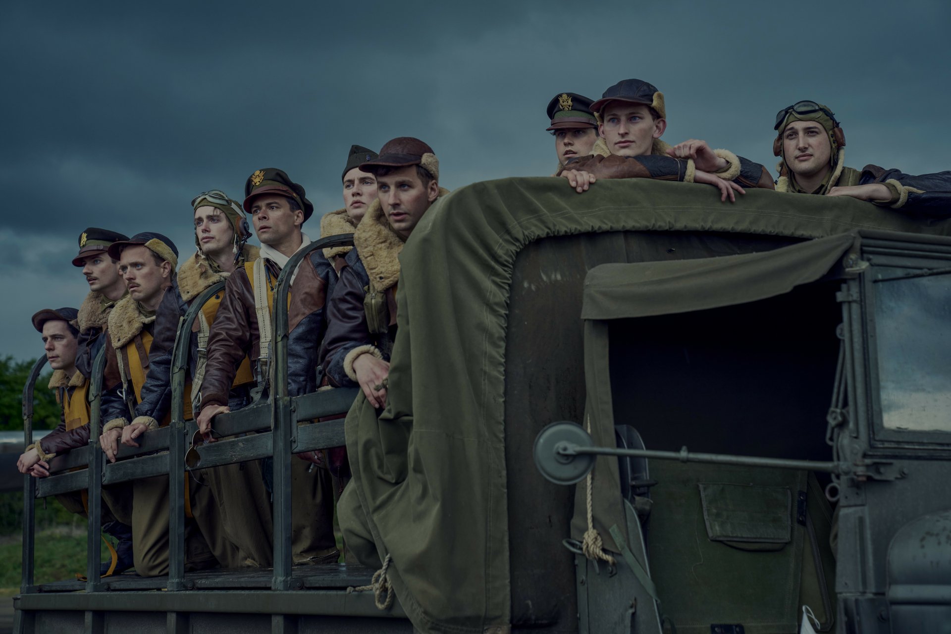 Group of actors portraying soldiers from 'Masters of the Air' TV show, standing in a military vehicle, featured in an HD desktop wallpaper.
