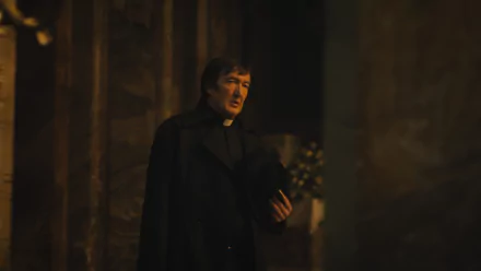 HD desktop wallpaper featuring a scene from The First Omen movie with a somber character in a clergy robe standing in a dimly lit setting, conveying a sense of mystery and suspense.