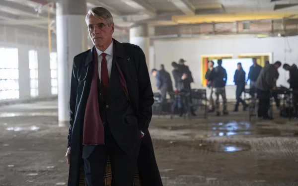 HD wallpaper of a scene from the movie 'The Beekeeper' featuring a distinguished man in a suit standing in a dimly lit, abandoned building with blurry figures in the background.