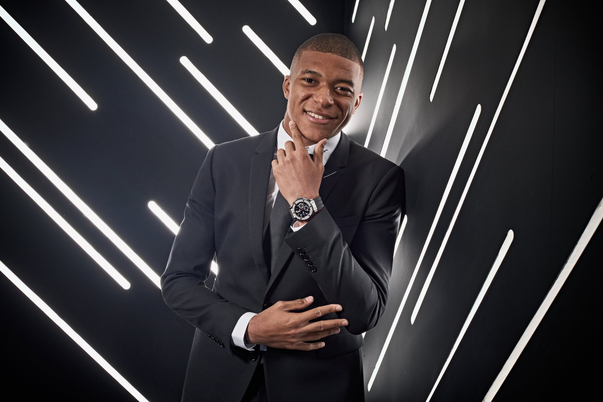 Stylish soccer star in a suit with a confident smile, posing for an HD desktop wallpaper against an abstract background of bright diagonal lines.