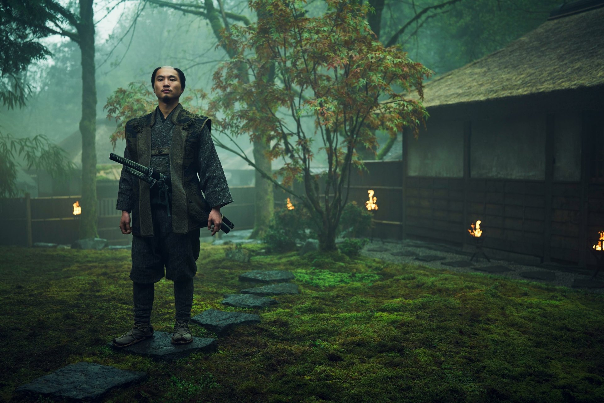 A scene from the TV show Shogun (2024), featuring a character in traditional attire standing in a misty, moss-covered landscape near a rustic, wooden structure.