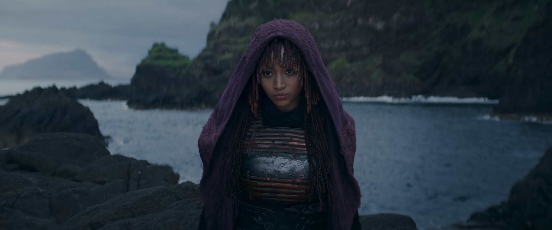 HD wallpaper of a mysterious character from 'The Acolyte' TV show (2024) standing on rocky shoreline with dramatic seascape in the background.