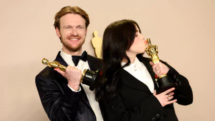 Billie Eilish and a smiling companion celebrate at the Academy Awards, each proudly holding an Oscar trophy. This vibrant HD desktop wallpaper captures their joyous moment.