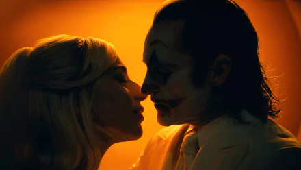 An HD desktop wallpaper showing a silhouetted man dressed as the Joker with white face paint and makeup facing a woman with blonde hair, both in profile with their faces close together, set against a warm, orange background.