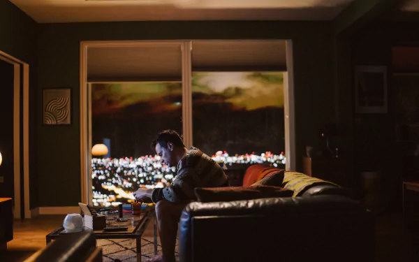 HD wallpaper featuring a contemplative scene from All of Us Are Strangers with a person sitting by the window overlooking city lights.