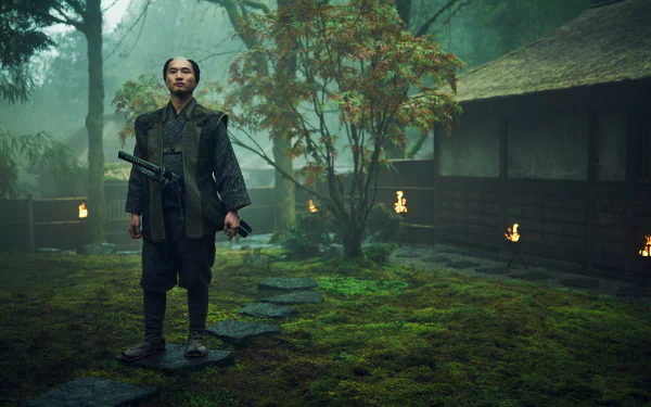 A scene from the TV show Shogun (2024), featuring a character in traditional attire standing in a misty, moss-covered landscape near a rustic, wooden structure.