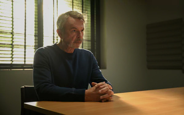 A character from the TV show Apples Never Fall sits thoughtfully at a table, illuminated by soft light, creating a dramatic atmosphere for a desktop wallpaper background.