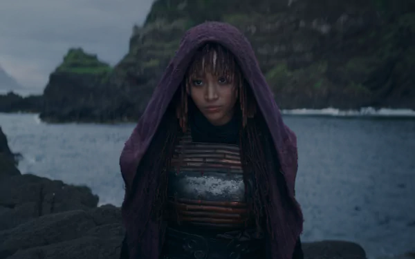 HD wallpaper of a mysterious character from 'The Acolyte' TV show (2024) standing on rocky shoreline with dramatic seascape in the background.