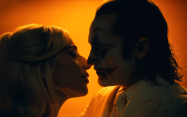 An HD desktop wallpaper showing a silhouetted man dressed as the Joker with white face paint and makeup facing a woman with blonde hair, both in profile with their faces close together, set against a warm, orange background.
