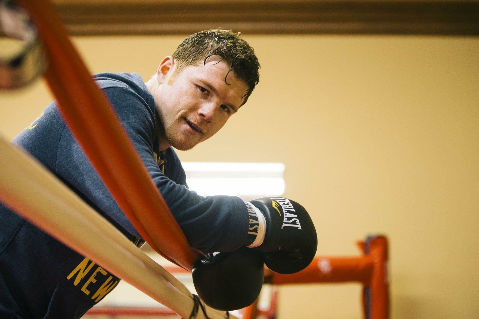 HD wallpaper of a male boxer in a gym, leaning on the ropes of a boxing ring, wearing gloves and a hoodie, focused and ready to train.
