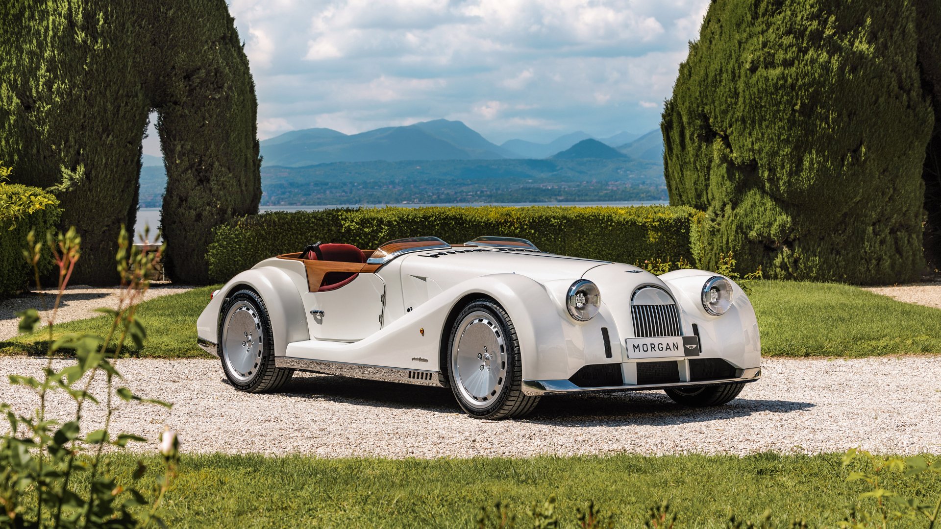 A stunning HD desktop wallpaper featuring a classic white Morgan Midsummer car, set against a picturesque background with lush greenery and distant mountains.