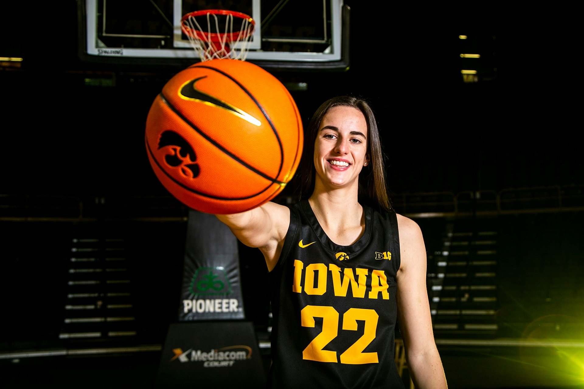 A high-definition desktop wallpaper featuring Caitlin Clark in an Iowa jersey holding a basketball with a confident smile. The background showcases a basketball hoop and a partially lit stadium.