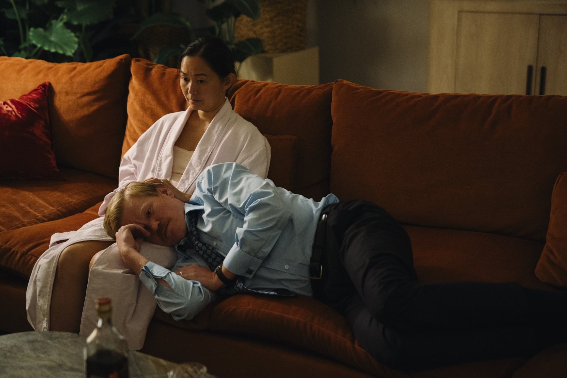 HD desktop wallpaper featuring a scene from the movie Kinds of Kindness, showing a woman sitting on a brown couch with a man resting his head on her lap, set in a cozy living room.