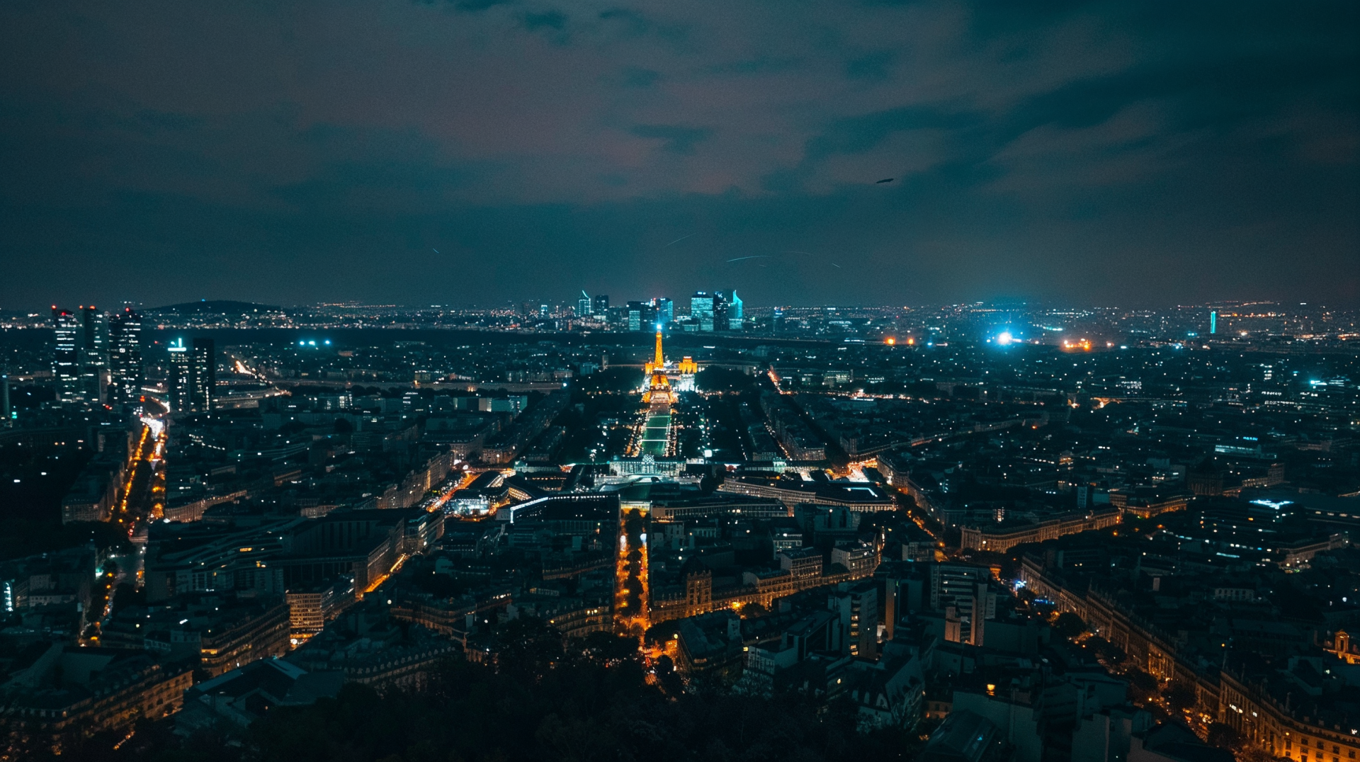 HD desktop wallpaper capturing a stunning night view of Paris with illuminated streets and the Eiffel Tower glowing in the center under a dark, cloudy sky.