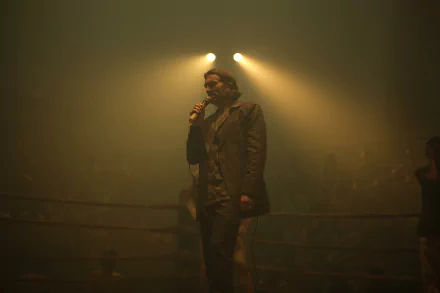 HD wallpaper featuring a scene from Monkey Man (2024) with a man in a trench coat standing in a foggy, dimly lit ring, spotlight overhead, addressing a crowd.