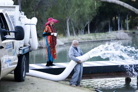 HD desktop wallpaper from the TV show Hacks featuring a whimsical scene with a character in a colorful outfit and another character by a pond, interacting with a hose that's expelling water.