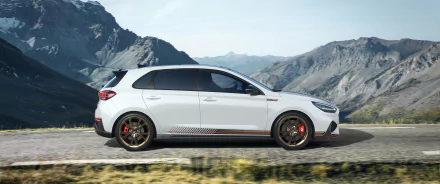 HD desktop wallpaper showcasing a white Hyundai i30 against a backdrop of scenic mountains and a clear sky. The car is seen from the side, highlighting its sleek design and detailed features.