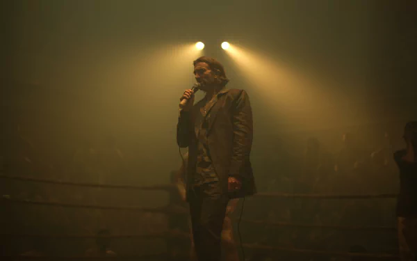 HD wallpaper featuring a scene from Monkey Man (2024) with a man in a trench coat standing in a foggy, dimly lit ring, spotlight overhead, addressing a crowd.