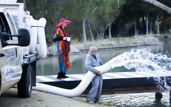 HD desktop wallpaper from the TV show Hacks featuring a whimsical scene with a character in a colorful outfit and another character by a pond, interacting with a hose that's expelling water.