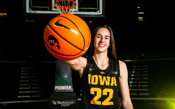 A high-definition desktop wallpaper featuring Caitlin Clark in an Iowa jersey holding a basketball with a confident smile. The background showcases a basketball hoop and a partially lit stadium.