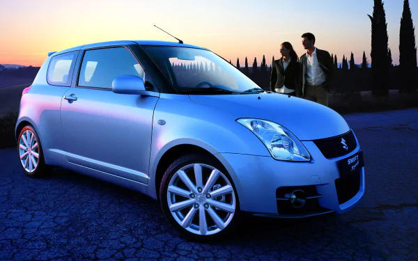 HD wallpaper showcasing a Suzuki Swift Sport car at sunset, with a couple in the background and a scenic landscape featuring tall trees.