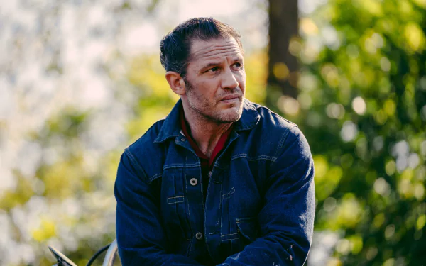 HD desktop wallpaper from the movie The Bikeriders, featuring Tom Hardy in a denim jacket, set against a natural blurred background.