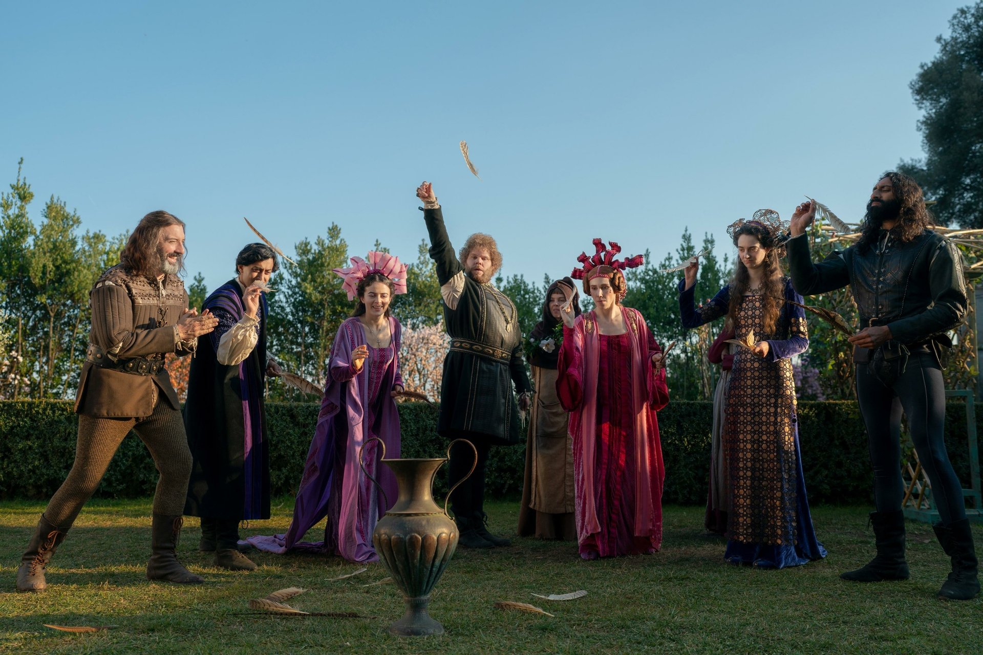 A vibrant scene from the TV show The Decameron, featuring a group of characters in historic costumes, joyfully interacting in a lush outdoor setting. مناسب كشاشة خلفية بدقة عالية.