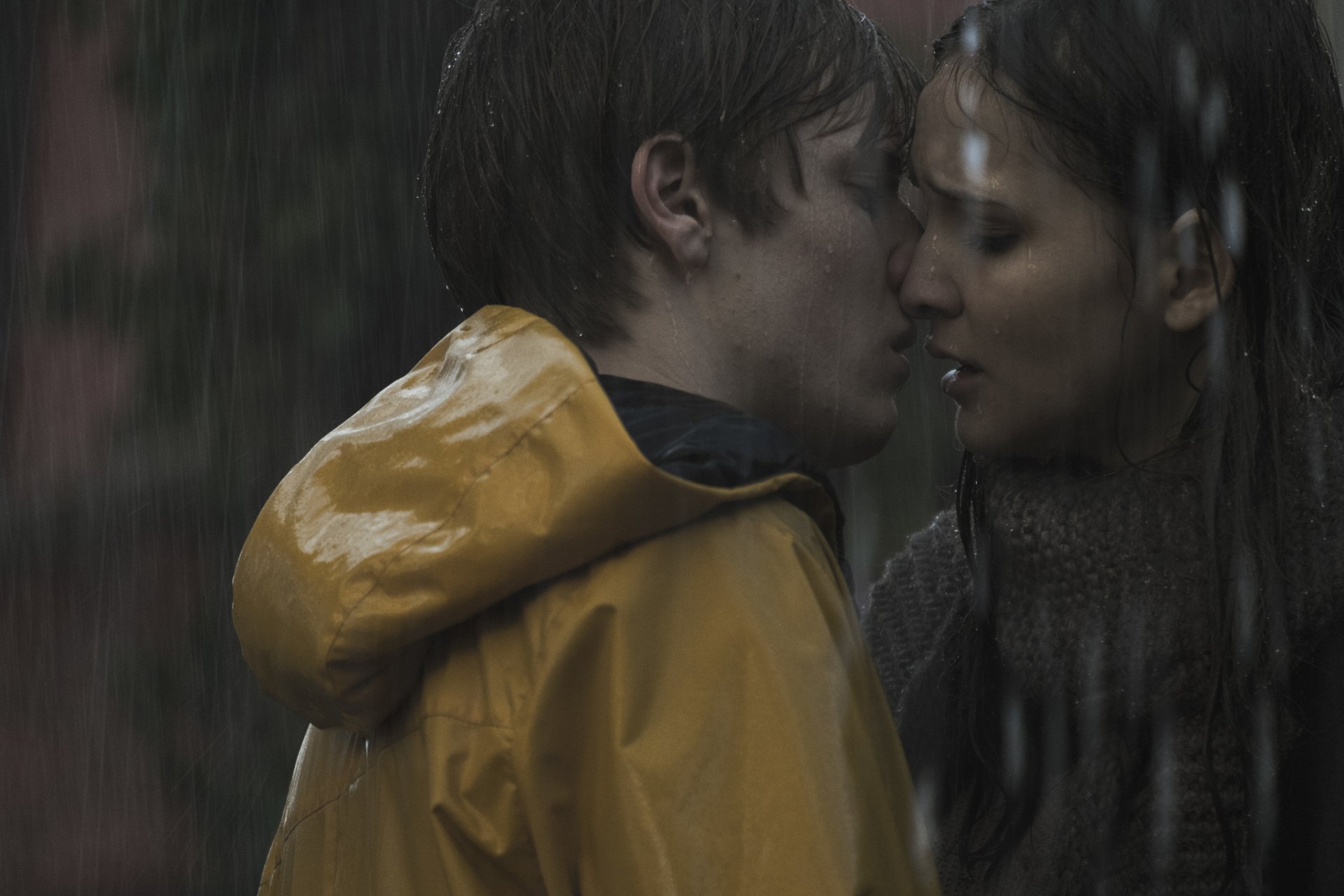 A captivating scene from the TV show Dark, featuring Louis Hofmann and Lisa Vicari sharing a tender moment in the rain, set against a dramatic backdrop. HD wallpaper.